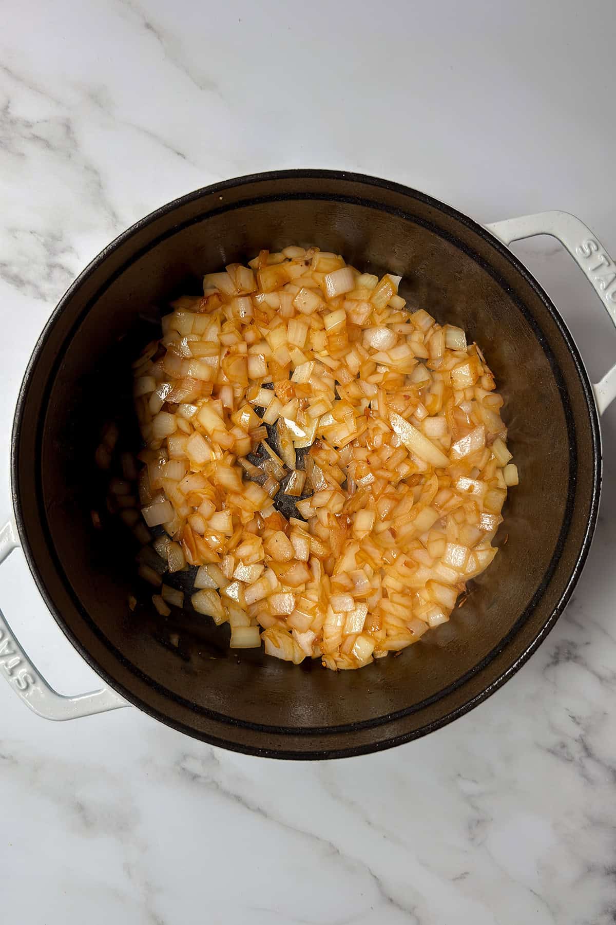 Diced onions are sautéing and turning golden brown in a black pot on a marble countertop, forming the flavorful base for a classic Irish Stew.