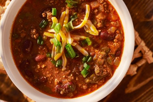 A bowl of hearty turkey chili, topped with shredded cheese and chopped green onions, sits on a woven mat. Next to it is a red and white checkered cloth. Two glass shakers are visible at the top of the image.