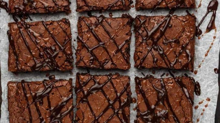 Nine square caramel brownies with a glossy drizzle of chocolate on top, arranged on parchment paper. Two antique-style dessert servers are placed below the brownies, adding a rustic touch.