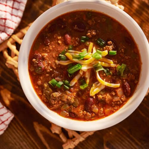 A bowl of turkey chili topped with shredded cheese and chopped green onions sits on a wooden table next to a red and white checkered towel and salt and pepper shakers.