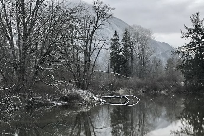 A serene winter scene with a calm river reflecting bare trees and a snow-dusted mountain in the background exudes an air of quiet kindness. Overcast skies add a tranquil, subdued atmosphere to the landscape.