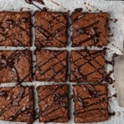 Nine caramel brownies drizzled with chocolate sauce are beautifully arranged on parchment paper. Two vintage silver utensils rest elegantly on the right side.