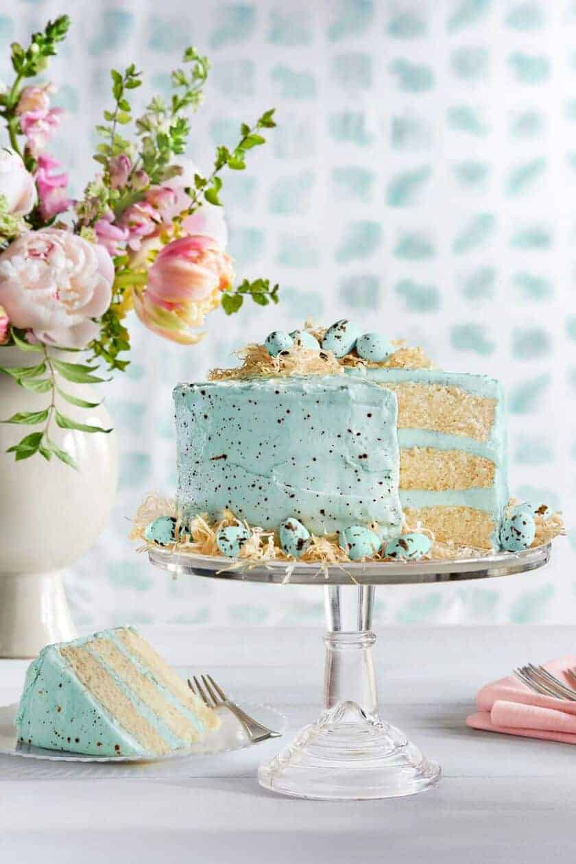 A light blue speckled Easter cake with three layers is adorned with small blue eggs and sits on a glass cake stand. A slice is cut and placed nearby. In the background, a vase with pink and white flowers completes the bright, fresh setting, perfect for trying new Easter cake recipes.