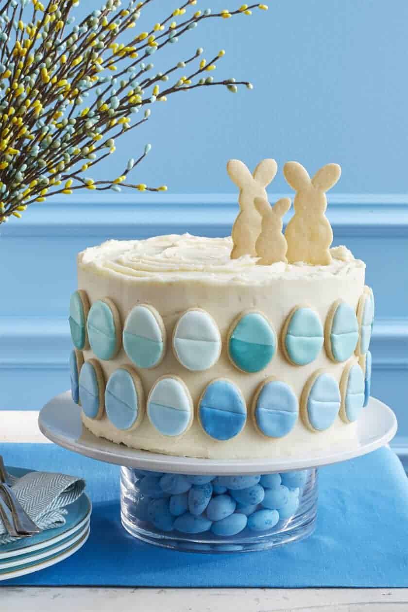 A decorated cake with white frosting and blue ombre egg-shaped cookies on the side. Two rabbit-shaped cookies hop atop this Easter delight on a clear stand filled with blue candies, next to a vase with branches and a stack of napkins.