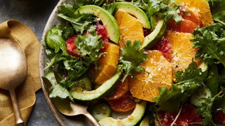 A white plate of strawberry spinach salad with pecans and crumbled cheese sits on a white napkin. Wooden serving utensils rest in the salad, and a small jar of dressing is nearby on a white wooden surface.