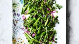 A rectangular plate with a colorful floral border holds a fresh spring salad for Easter, featuring green peas, snap peas, and pea shoots mixed with sliced red onions. This delightful Easter dish is beautifully arranged on a white marble surface.