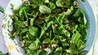 A fresh Easter salad with mixed greens, mint leaves, blue cheese crumbles, and toasted seeds is served in a decorative floral-patterned bowl on a floral tablecloth.