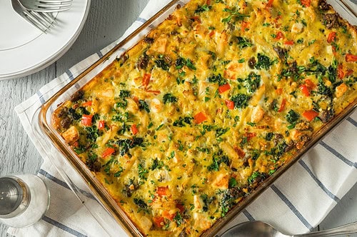 A casserole dish filled with a vibrant baked strata, showcasing spinach, red bell pepper, and hearty chunks of bread intertwined with a delicious sausage, egg, and kale breakfast blend. It rests on a blue and white striped cloth with plates, a fork, and a pepper grinder close by.