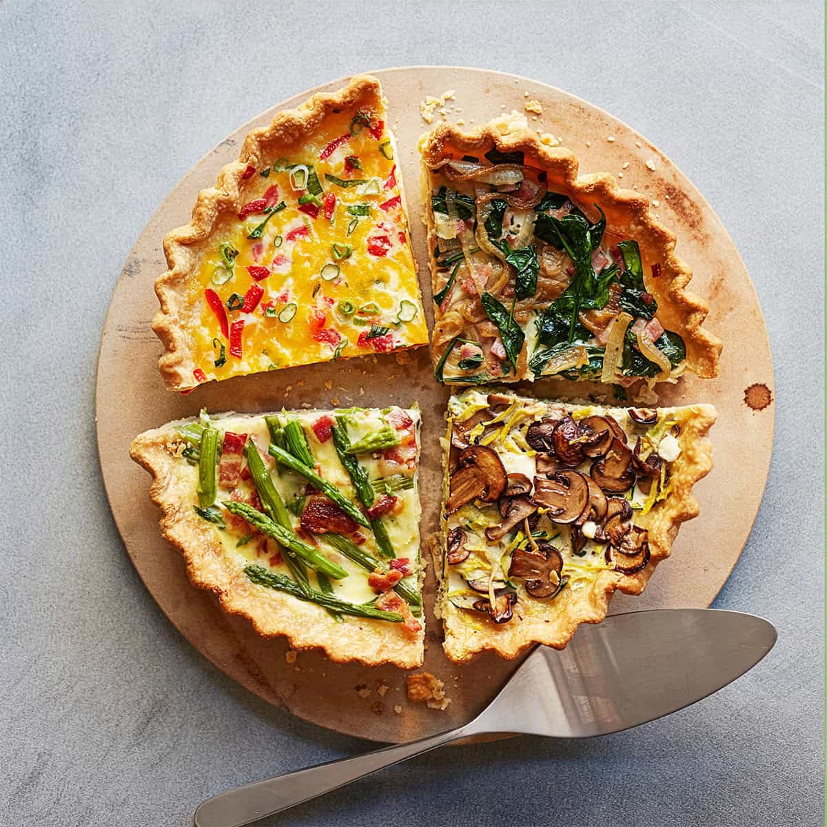 Perfect for brunch, this quiche is divided into four quarters with unique toppings: corn and peppers, spinach and onions, asparagus and bacon, and mushrooms with herbs. A metal pie server rests beside the quiche.