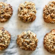 Six oatmeal chocolate coconut cookies are arranged in two rows on a light marble surface. The cookies boast a golden-brown hue and textured appearance, hinting at their chewy consistency.
