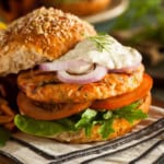 A salmon burger with lettuce, tomato, red onion, and a dollop of sauce on a whole grain bun, served on a striped napkin.