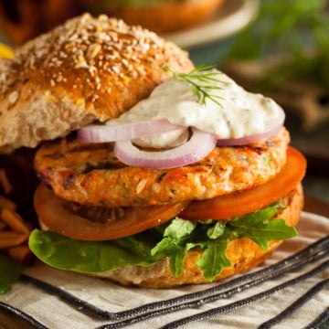 A salmon burger with lettuce, tomato, red onion, and a dollop of sauce on a whole grain bun, served on a striped napkin.