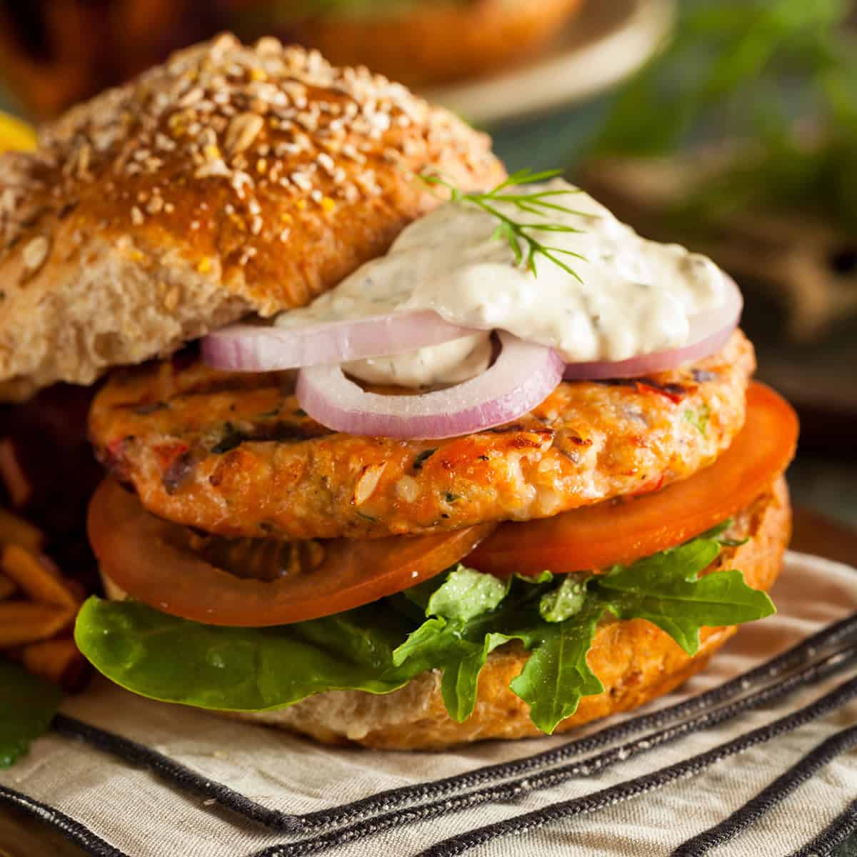 A salmon burger with lettuce, tomato, red onion, and a dollop of sauce on a whole grain bun, served on a striped napkin.