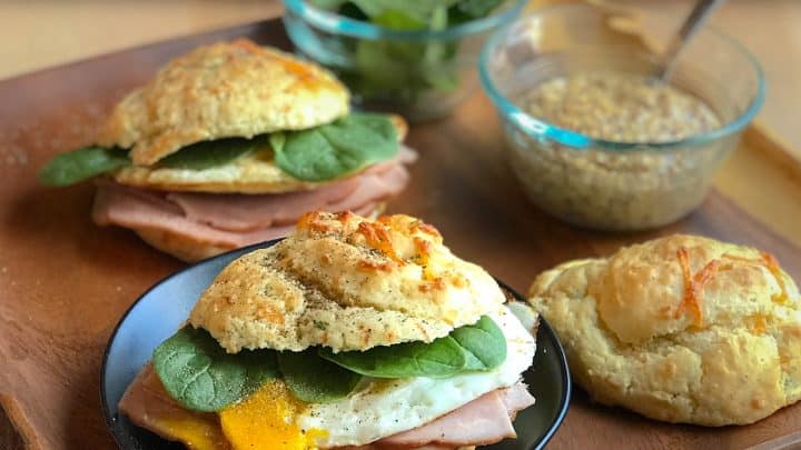 A pink frosted layer cake on a white stand, decorated with raspberries, pink meringues, and small pink flowers—perfect for a Mother's Day Breakfast—set on a pink wooden surface with plates and utensils nearby.