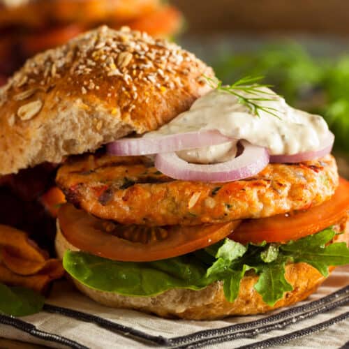 Enjoy our salmon burgers on a whole grain bun with lettuce, tomato, red onion slices, and creamy dill sauce, served alongside crispy vegetable chips.