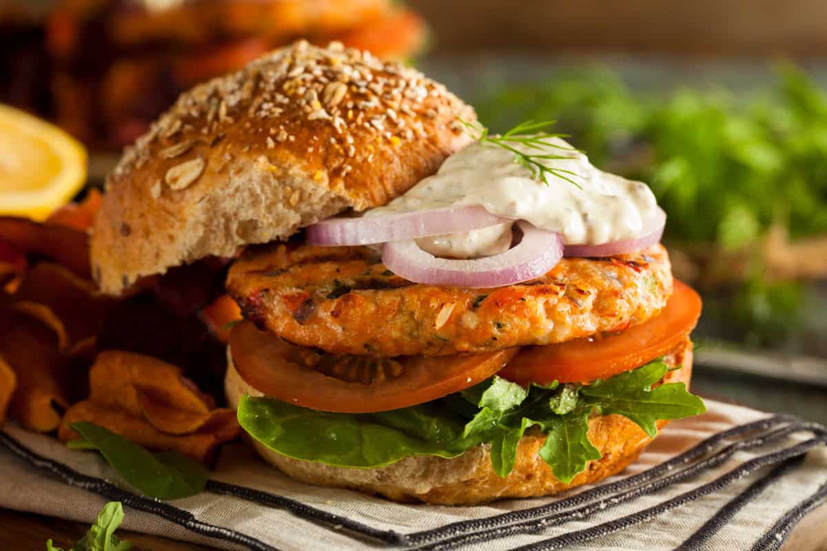 Enjoy our salmon burgers on a whole grain bun with lettuce, tomato, red onion slices, and creamy dill sauce, served alongside crispy vegetable chips.