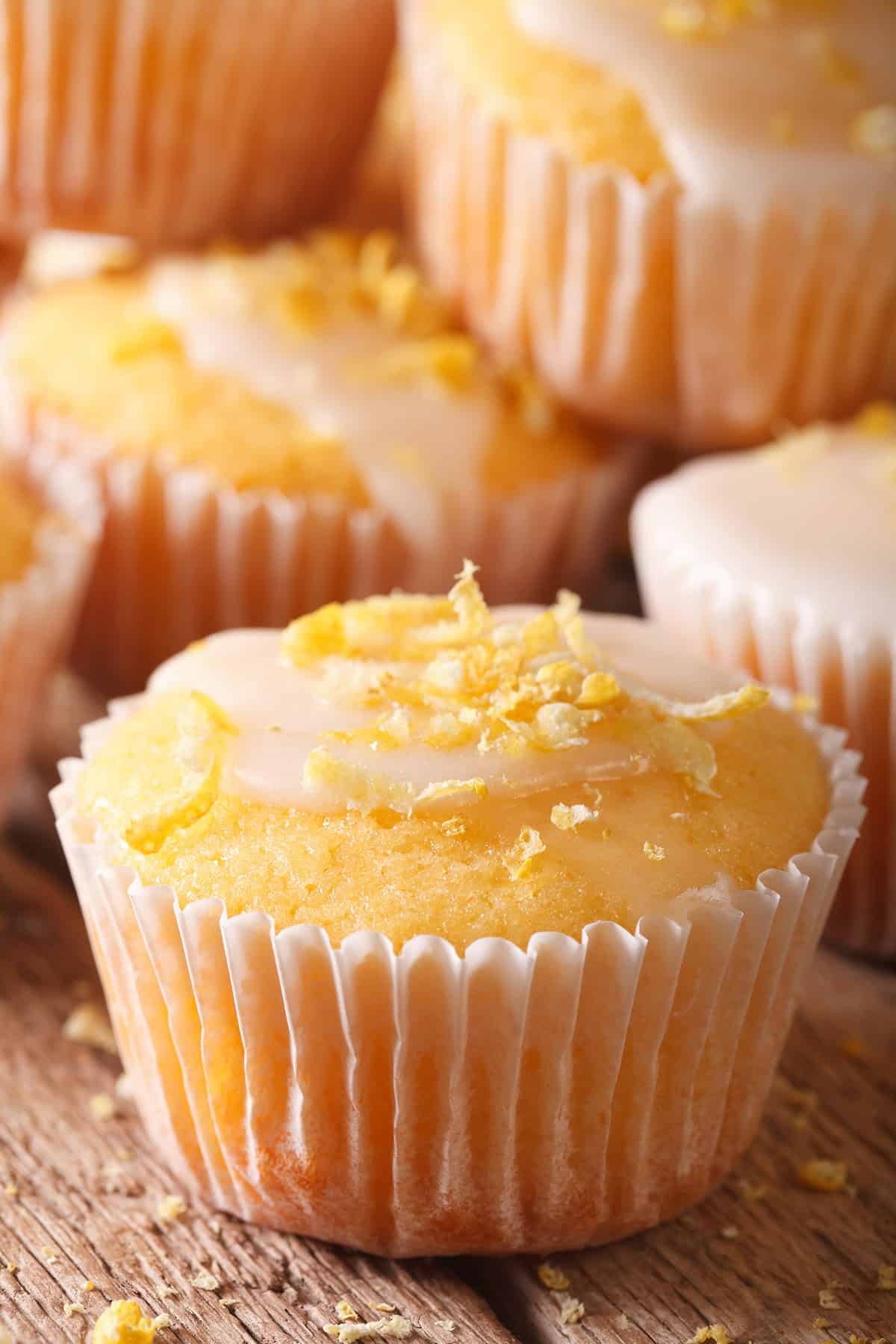 Close-up of greek yogurt lemon muffins with white icing and lemon zest on top, arranged on a wooden surface.