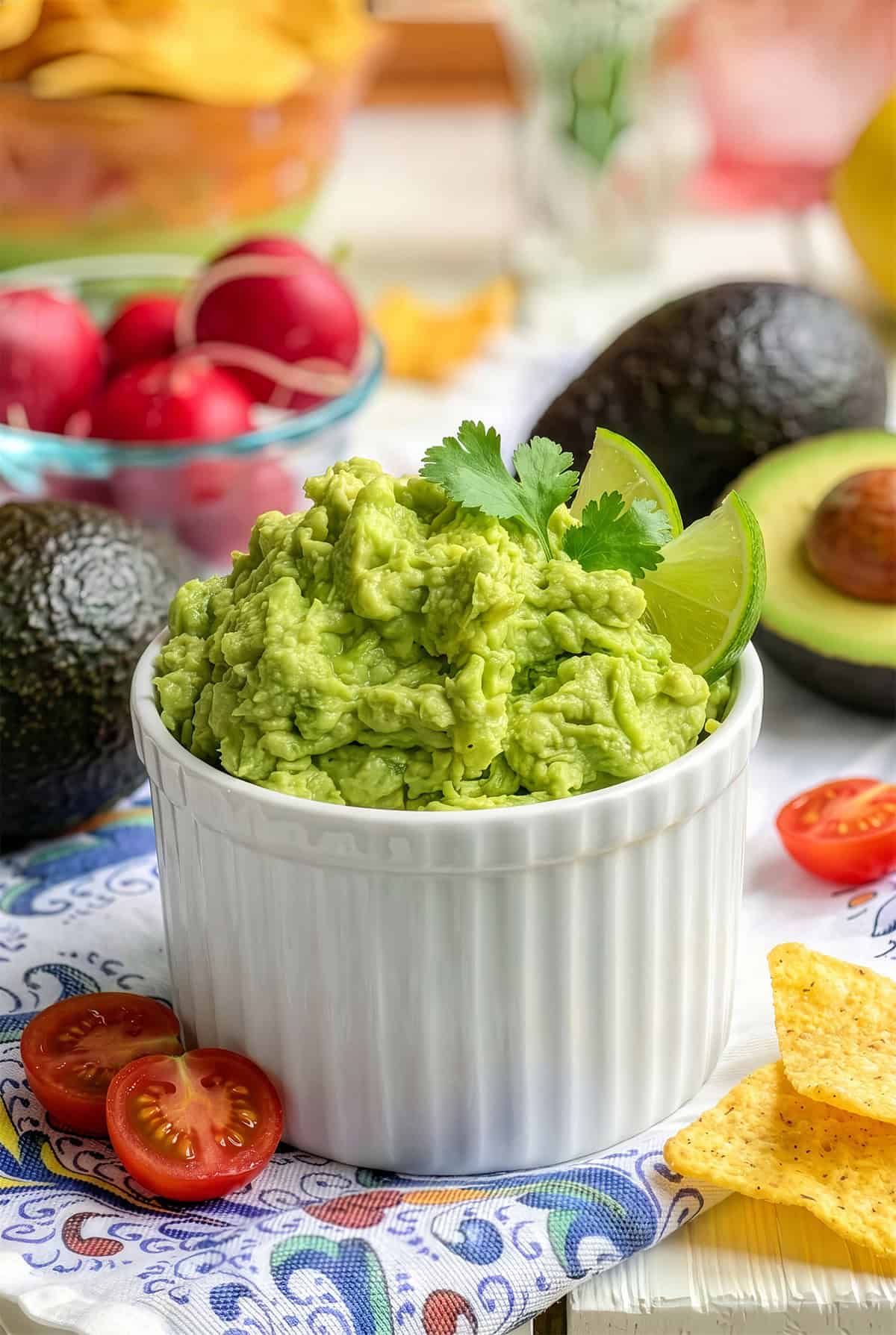 A white ramekin filled with creamy guacamole, garnished with lime wedges and cilantro, is surrounded by avocados, cherry tomatoes, radishes, and tortilla chips.