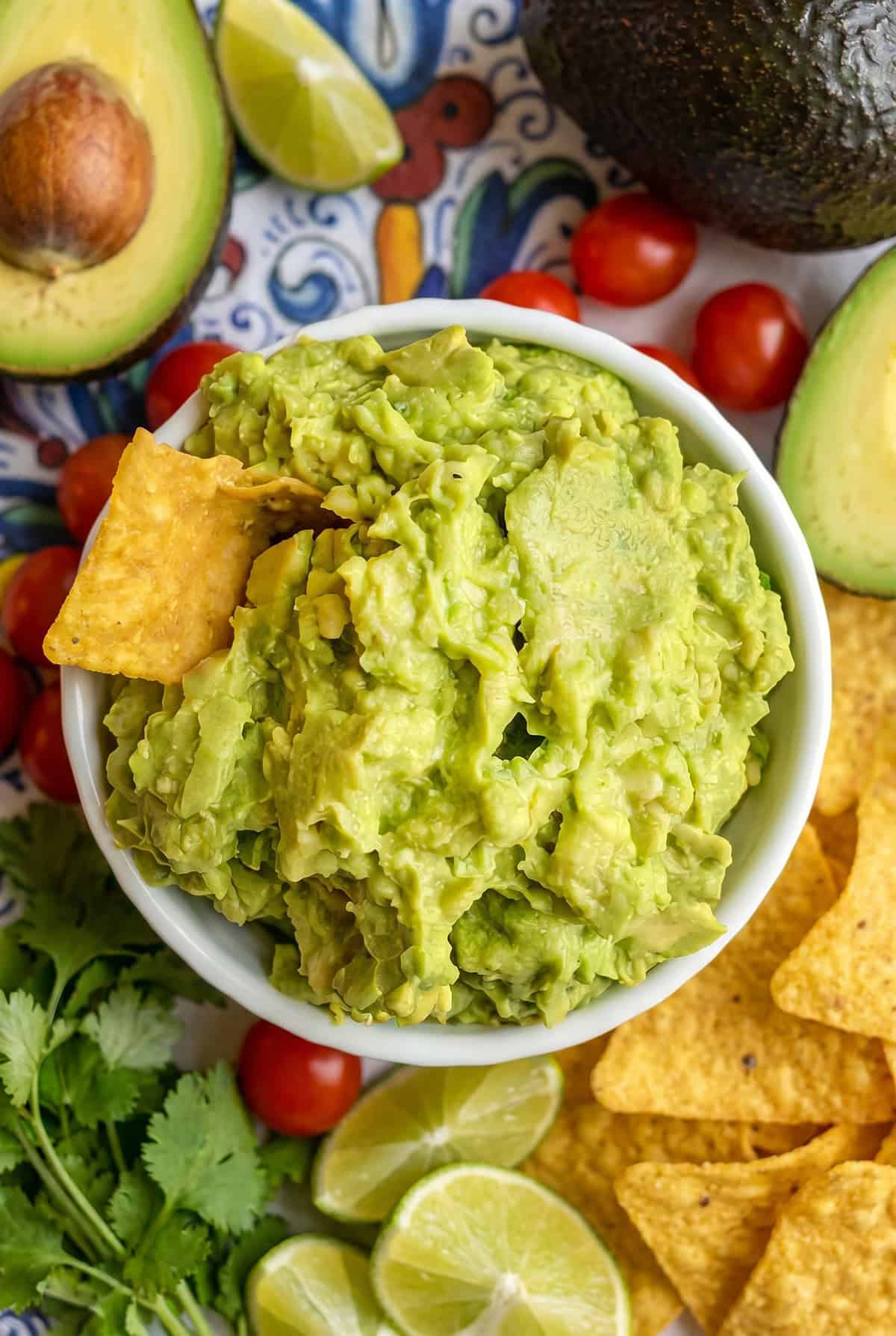 A bowl of fresh guacamole with a tortilla chip, surrounded by avocado halves, cherry tomatoes, lime wedges, cilantro, and extra tortilla chips.