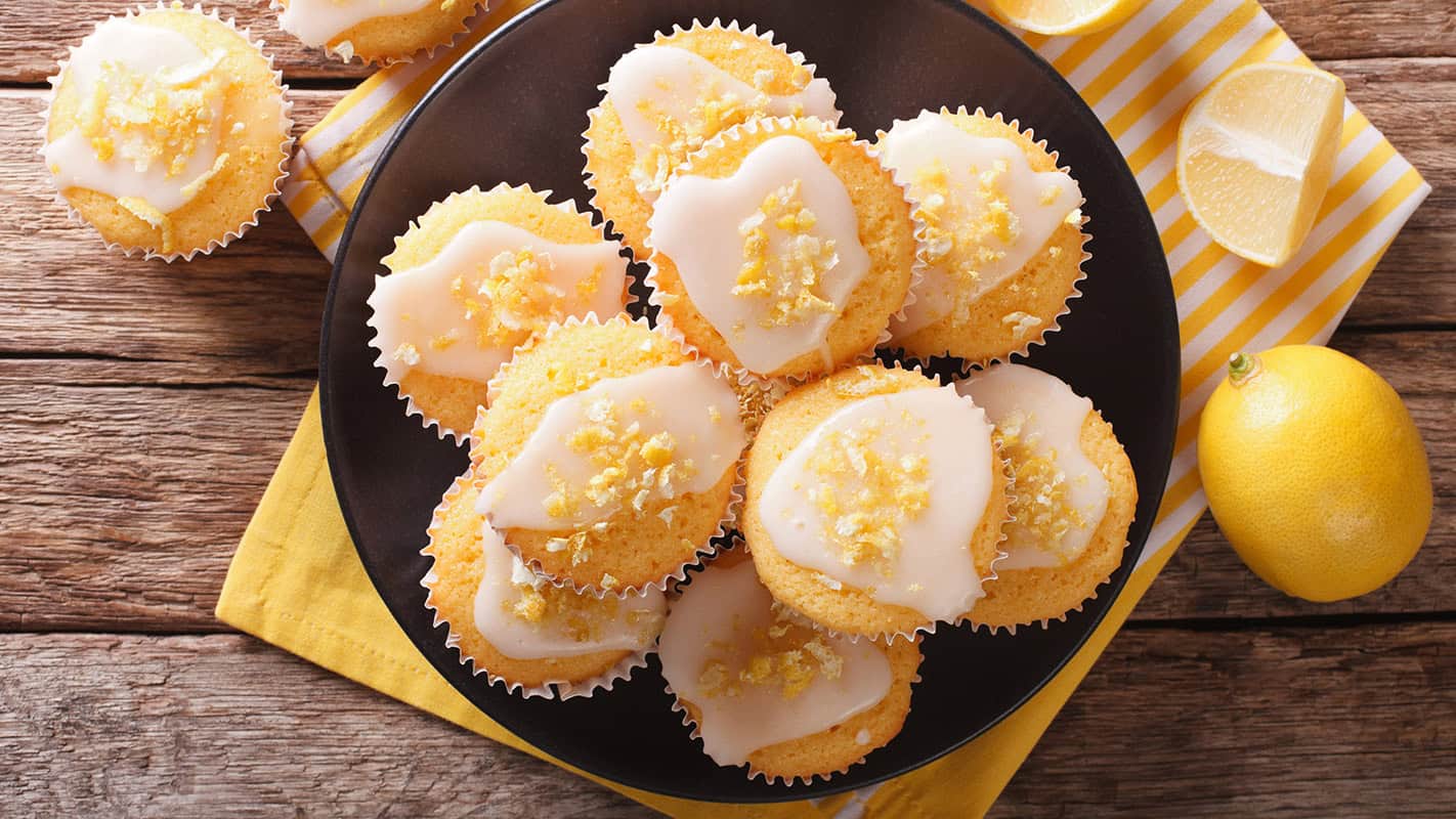 A plate of lemon yogurt muffins with white icing and lemon zest on top, surrounded by whole and sliced lemons on a rustic wooden table.