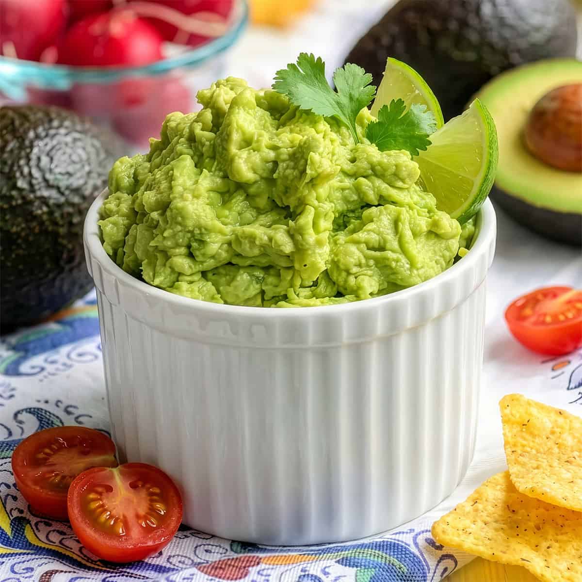 A white ramekin filled with fresh guacamole, garnished with lime and cilantro, surrounded by tomatoes, tortilla chips, avocados, and radishes.
