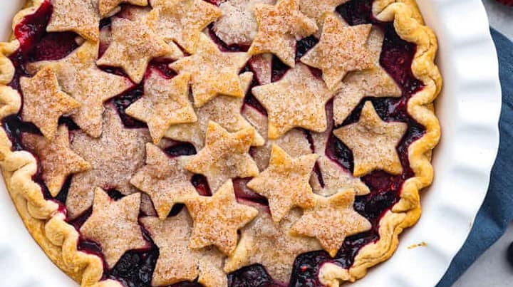 A baked berry pie in a white dish, topped with star-shaped crust pieces, surrounded by fresh strawberries and blueberries—perfect for red white and blue recipes on a light surface.