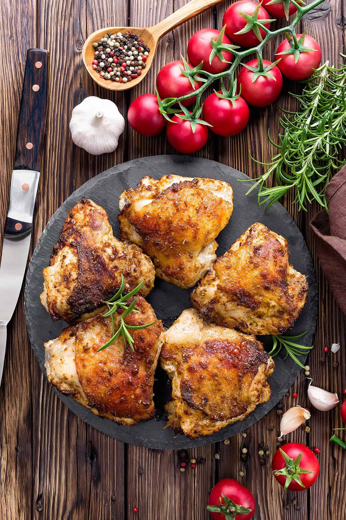Four pieces of Grilled Lemon and Herb Chicken Thighs on a dark plate, garnished with rosemary, surrounded by tomatoes, garlic, peppercorns, and a knife on a wooden table.