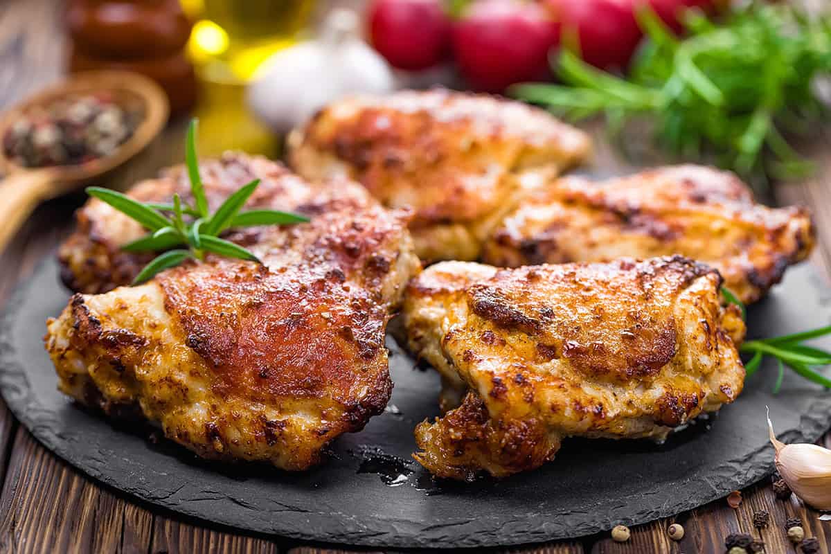 Five pieces of Grilled Lemon and Herb Chicken Thighs on a black slate plate, garnished with rosemary, with herbs and spices in the background.
