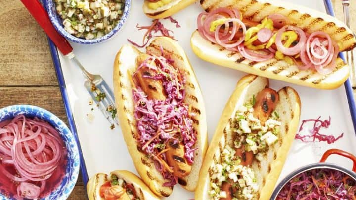 A tray of grilled hot dogs in buns topped with onions, slaw, and pickles—a classic Memorial Day food—served with pickled onions, extra slaw, a soda can, and a small American flag on a wooden table.