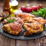 Grilled Lemon and Herb Chicken Thighs garnished with rosemary rest on a slate plate, surrounded by peppercorns, oil, garlic, and radishes on a rustic wooden table.