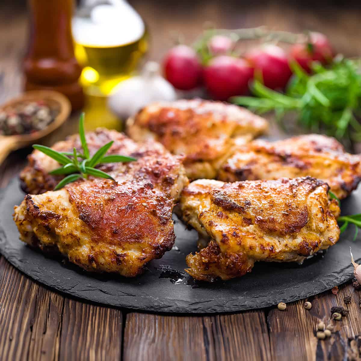 Grilled Lemon and Herb Chicken Thighs garnished with rosemary rest on a slate plate, surrounded by peppercorns, oil, garlic, and radishes on a rustic wooden table.