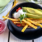 A black bowl brimming with chicken tortilla soup is garnished with avocado slices, sour cream, and tortilla strips. A small glass dish of pickled onions sits beside it, all arranged on a white wooden surface with a light green cloth nearby.