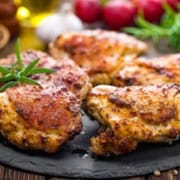 Grilled Lemon and Herb Chicken Thighs seasoned with pepper, arranged on a round slate platter. In the background, fresh rosemary, radishes, a garlic bulb, and a wooden spoon with peppercorns are visible.