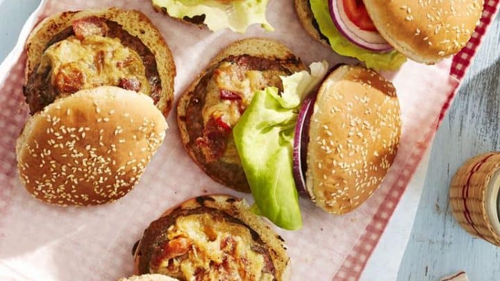 Six cheeseburgers with lettuce, tomato, and onion on sesame seed buns are arranged on a tray lined with pink checkered paper—perfect for your Memorial Day cookout recipes.