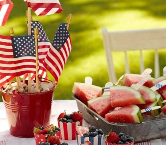 A table set for a 4th of July cookout displays a bucket of American flags, a tray of watermelon slices with tiny flags, and berry-filled cups in red, white, and blue starry patterns. A chair sits peacefully in the background on the grass.