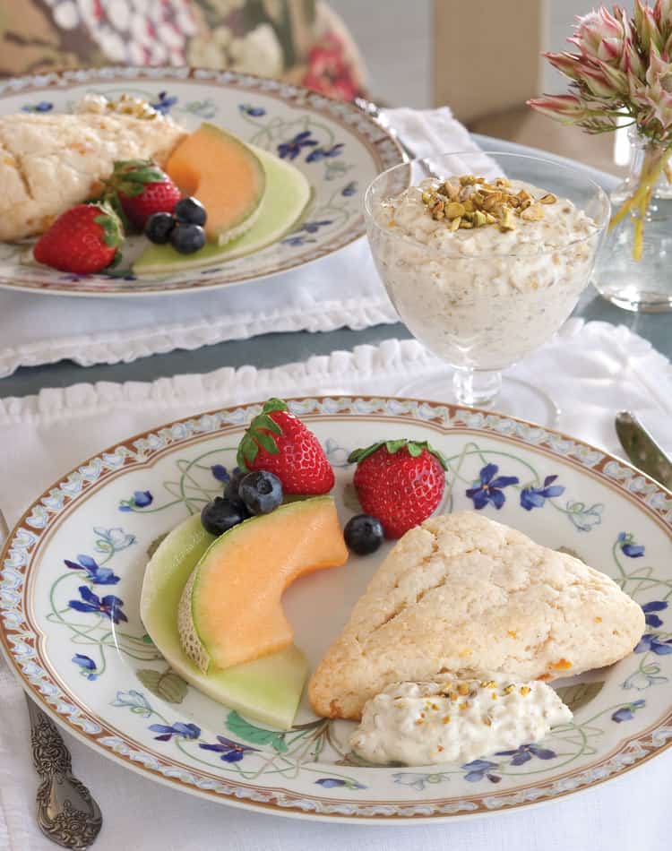 A plate with afternoon tea scones, cantaloupe, honeydew slices, strawberries, blueberries, and a dollop of cream. Next to it is a glass of muesli topped with nuts.
