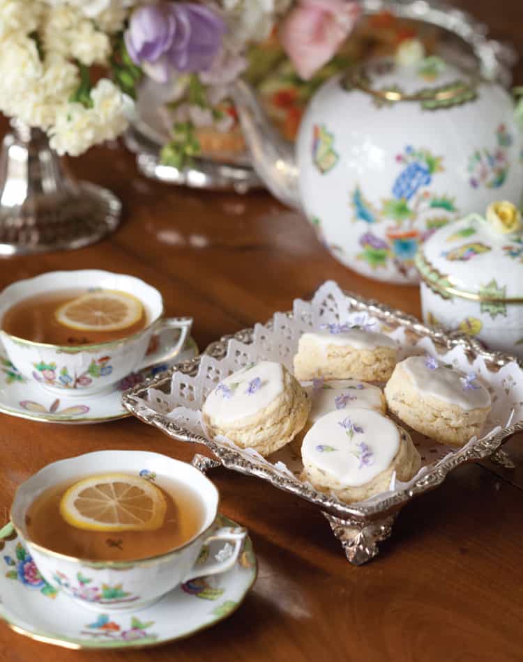 Two cups of tea with lemon slices and a tray of frosted afternoon tea scones are arranged on a wooden table with a floral porcelain teapot, sugar bowl, and fresh flowers in the background.