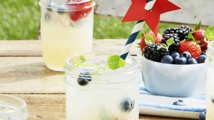 Close-up of lemonades in glass jars, garnished with berries and mint, complete with red and blue star-shaped straw decorations perfect for a 4th of July cookout. A bowl of assorted berries sits on a wooden outdoor table, grass visible in the background.
