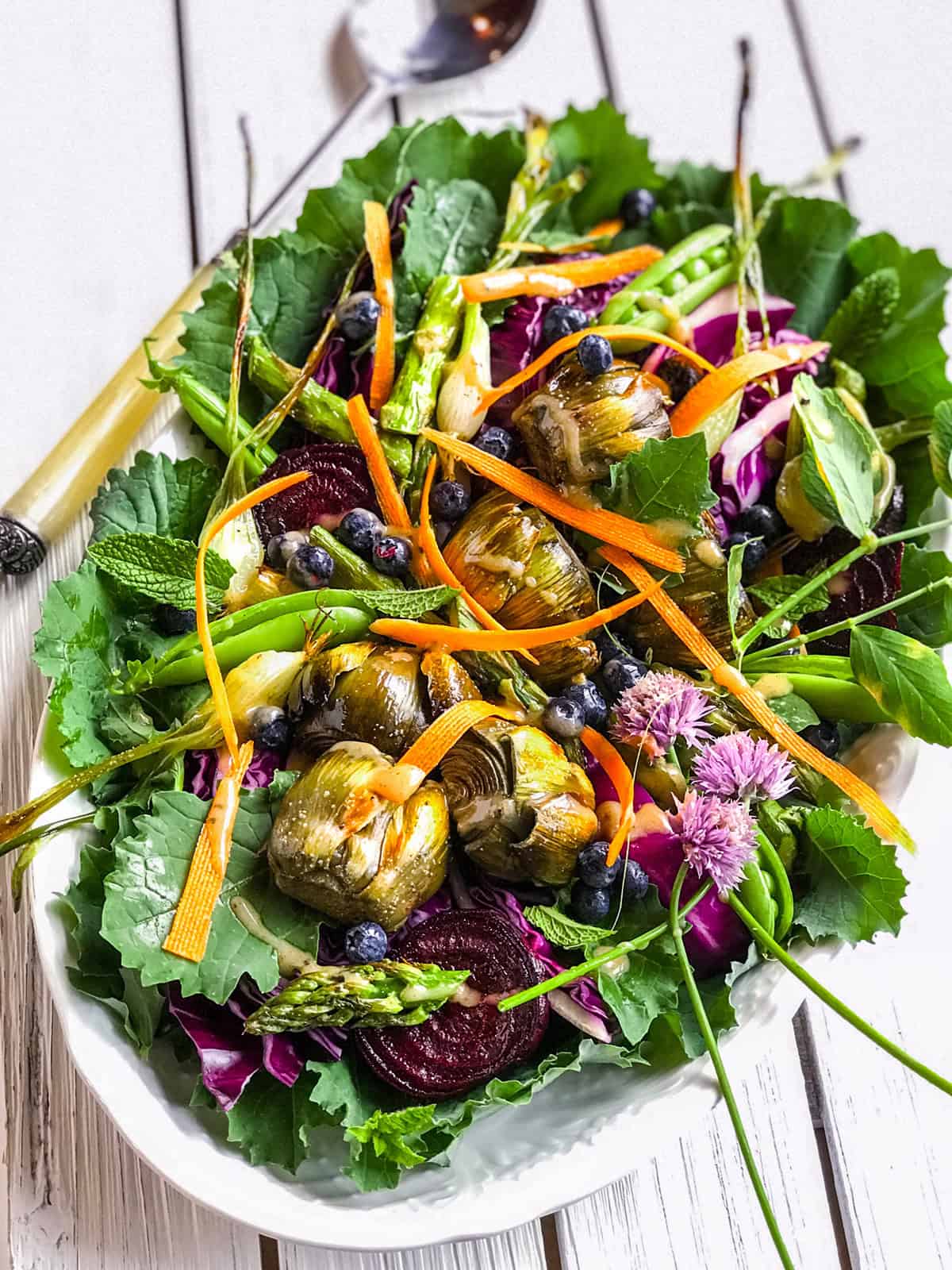 A colorful antipasto salad with leafy greens, grilled artichokes, asparagus, carrot ribbons, beet slices, blueberries, and edible flowers arranged beautifully on a white plate.