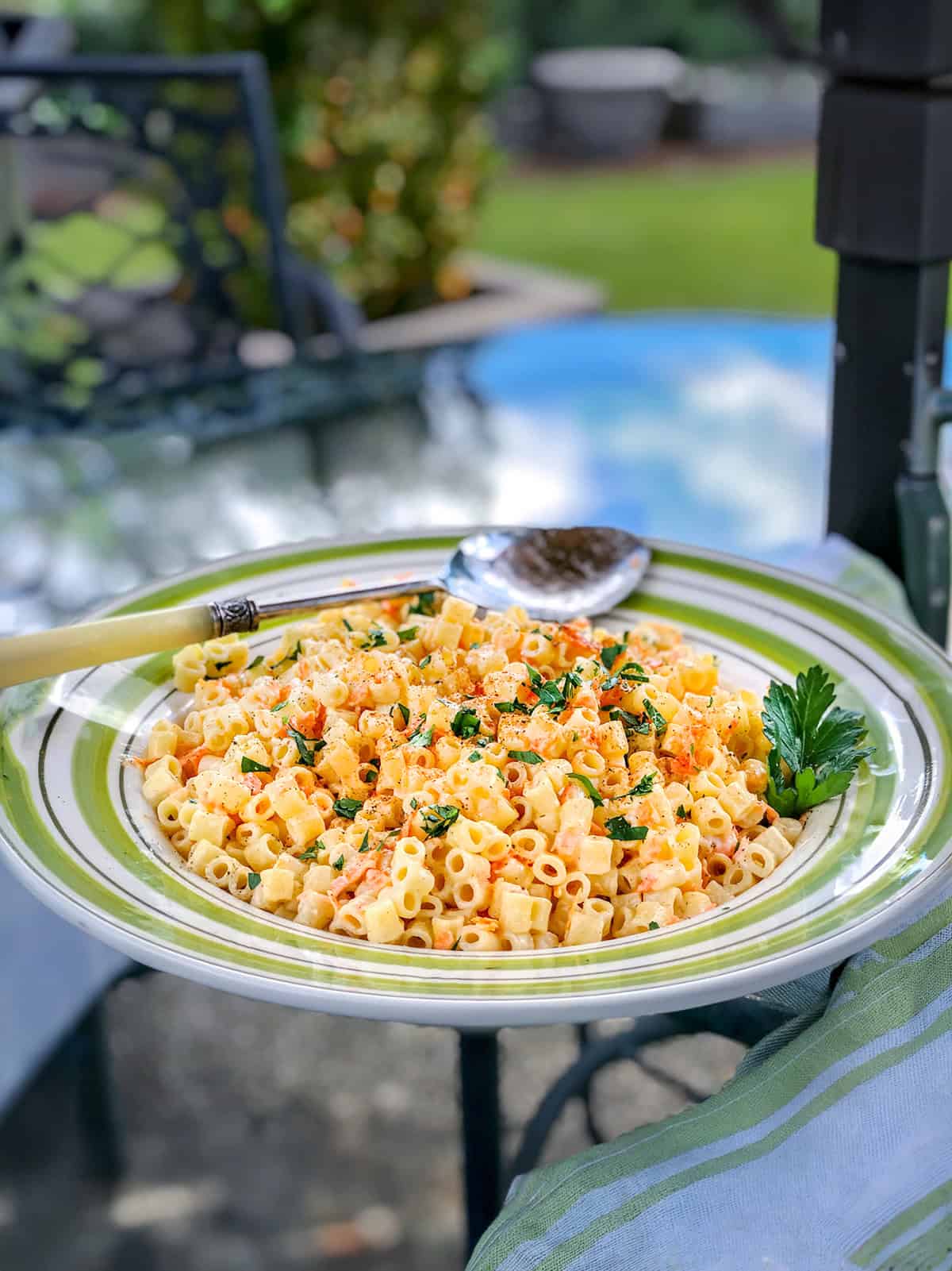 A plate of pasta salad with shredded carrots, small pasta tubes, diced vegetables, and herbs is garnished with parsley and sits on an outdoor table with a spoon beside it.