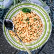 A bowl of pasta salad with shredded carrots, topped with chopped parsley and paprika, with a spoon resting on the side of the bowl.