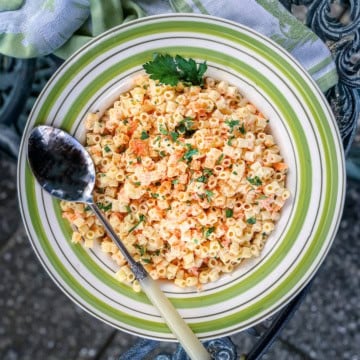 A bowl of pasta salad with shredded carrots, topped with chopped parsley and paprika, with a spoon resting on the side of the bowl.
