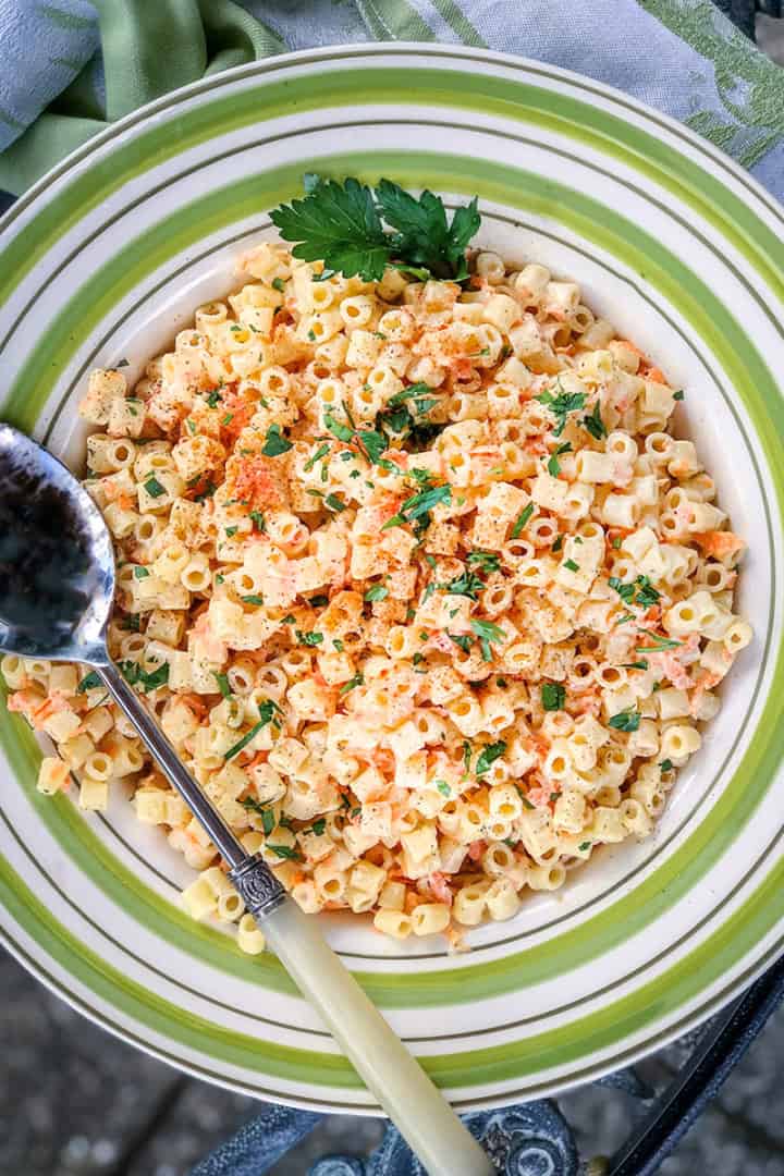 A bowl of pasta salad with shredded carrots, topped with chopped parsley and paprika, with a spoon resting on the side of the bowl.
