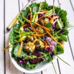 A vibrant antipasto salad platter featuring mixed greens, artichoke hearts, asparagus, carrot ribbons, beet slices, blueberries, and edible flowers on a white wooden table.