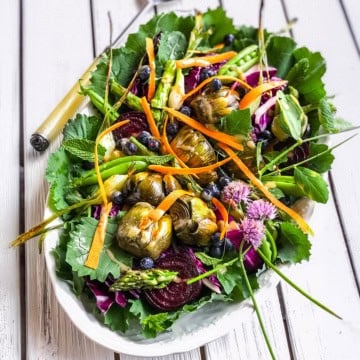 A vibrant antipasto salad platter featuring mixed greens, artichoke hearts, asparagus, carrot ribbons, beet slices, blueberries, and edible flowers on a white wooden table.