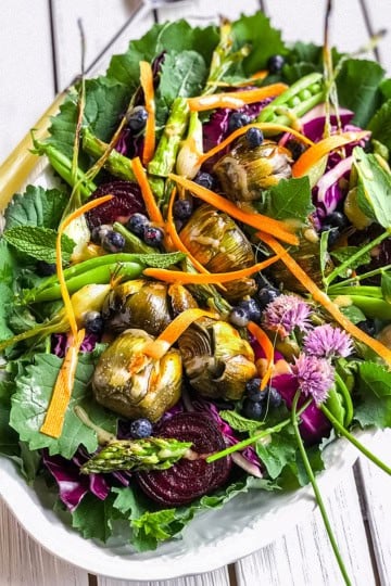 A vibrant antipasto salad platter featuring mixed greens, artichoke hearts, asparagus, carrot ribbons, beet slices, blueberries, and edible flowers on a white wooden table.