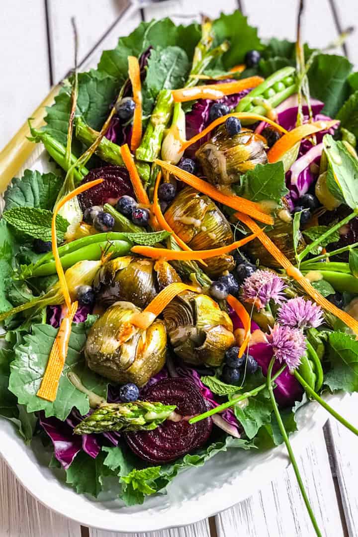A vibrant antipasto salad platter featuring mixed greens, artichoke hearts, asparagus, carrot ribbons, beet slices, blueberries, and edible flowers on a white wooden table.