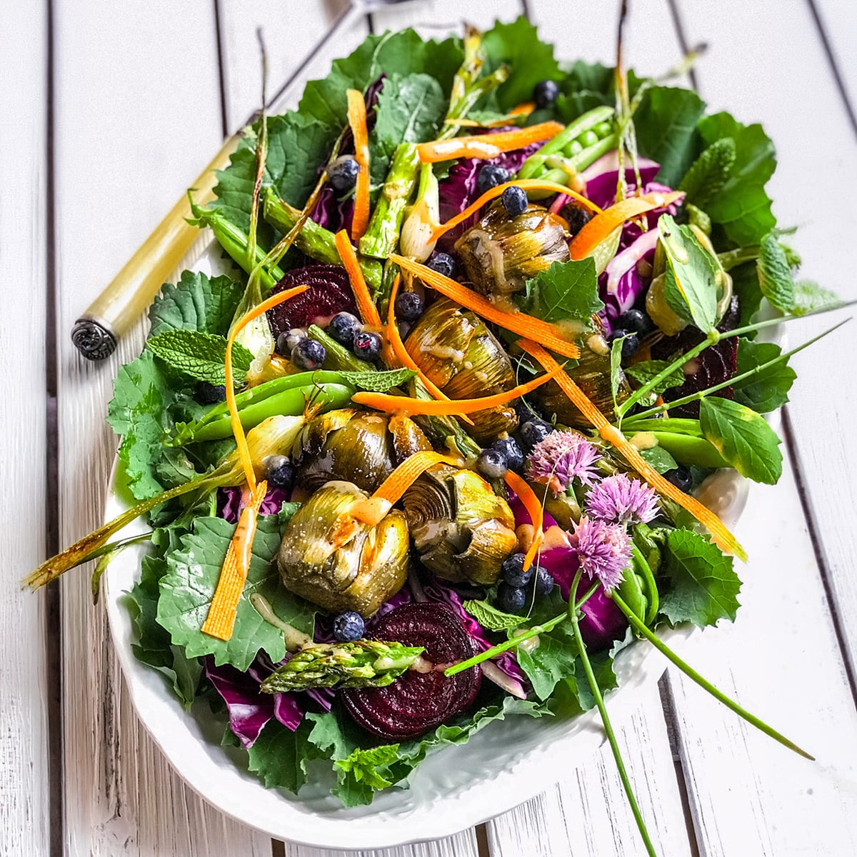 A vibrant antipasto salad platter featuring mixed greens, artichoke hearts, asparagus, carrot ribbons, beet slices, blueberries, and edible flowers on a white wooden table.