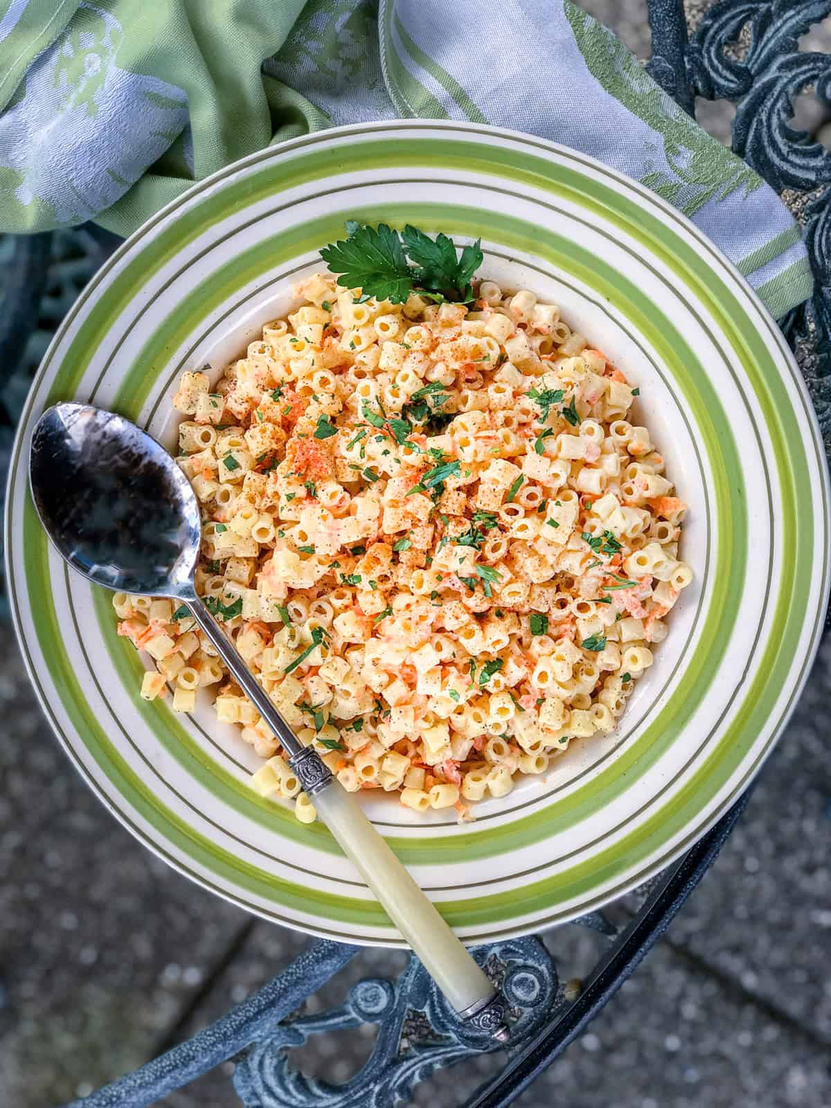 A bowl of creamy pasta salad with shredded carrots, topped with chopped herbs and paprika, served with a spoon on a green-striped plate.