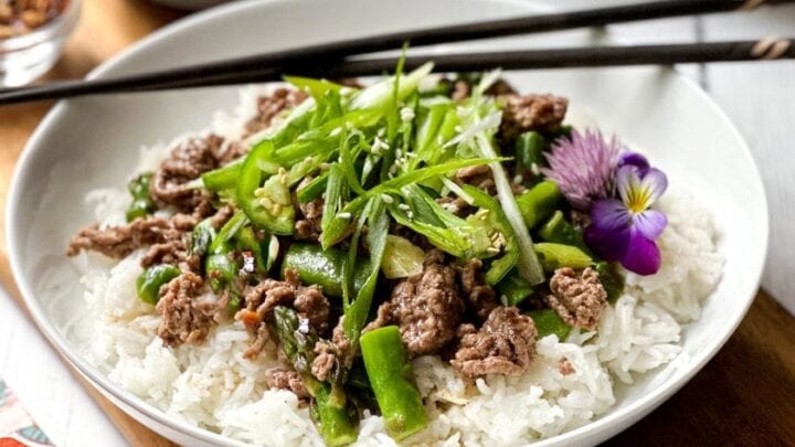 A bowl of white rice topped with Thai ginger beed and a garnish of purple edible flowers and shredded scallions.